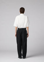 A person stands facing away on a plain light gray background, wearing the IM MEN “CLAY” loose white short-sleeve shirt tucked into high-waisted black tapered pants.