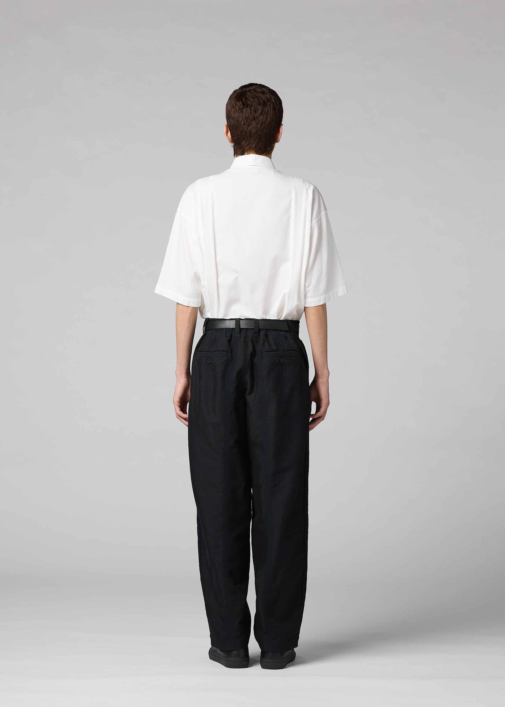 Zoomed view A person stands facing away on a plain light gray background, wearing the IM MEN “CLAY” loose white short-sleeve shirt tucked into high-waisted black tapered pants.