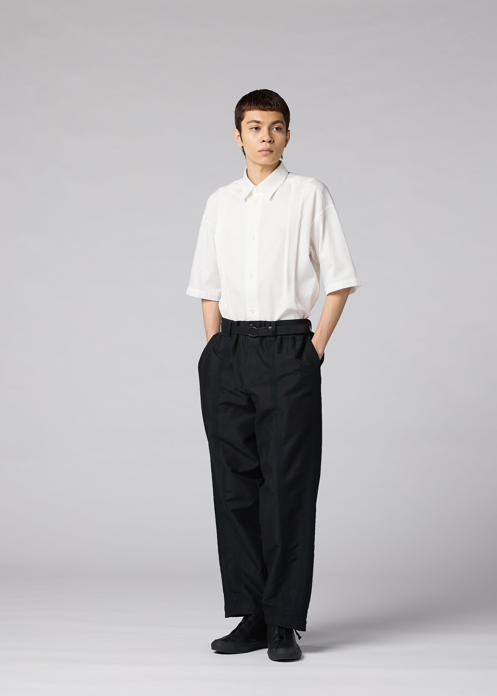 Zoomed view A person stands against a plain background, wearing IM MEN’s CLAY white short-sleeve button-up shirt crafted from sculptural fabric. The shirt is tucked into high-waisted, tapered black pants, paired with black shoes and hands in pockets.