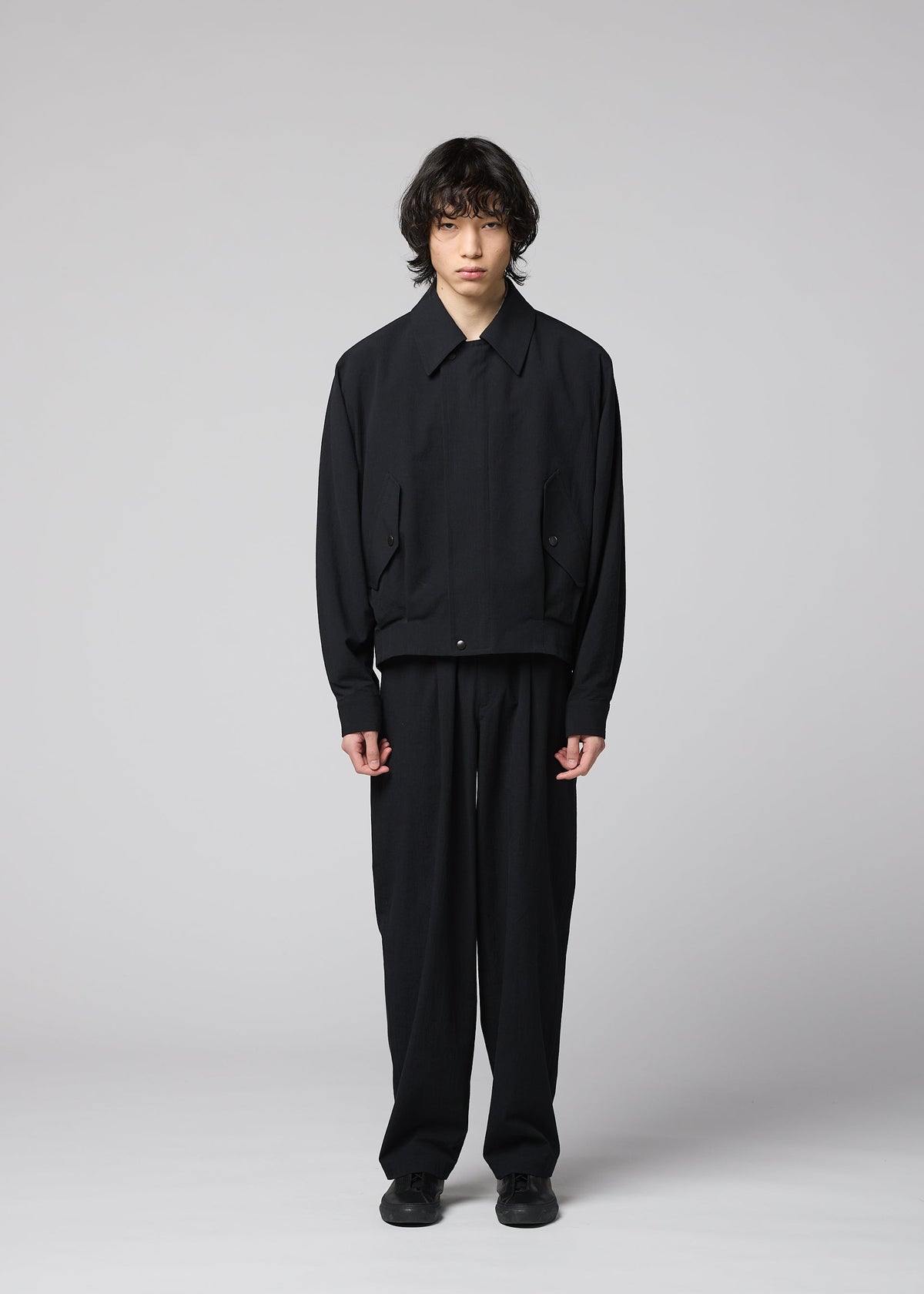 A person with medium-length dark hair stands against a plain light background, wearing the IM MEN COMFORT COTTON loose-fitting black jacket and straight pants crafted from high-twist organic cotton for a smooth feel, paired with black shoes, looking straight at the camera with a neutral expression. view 5 of 8