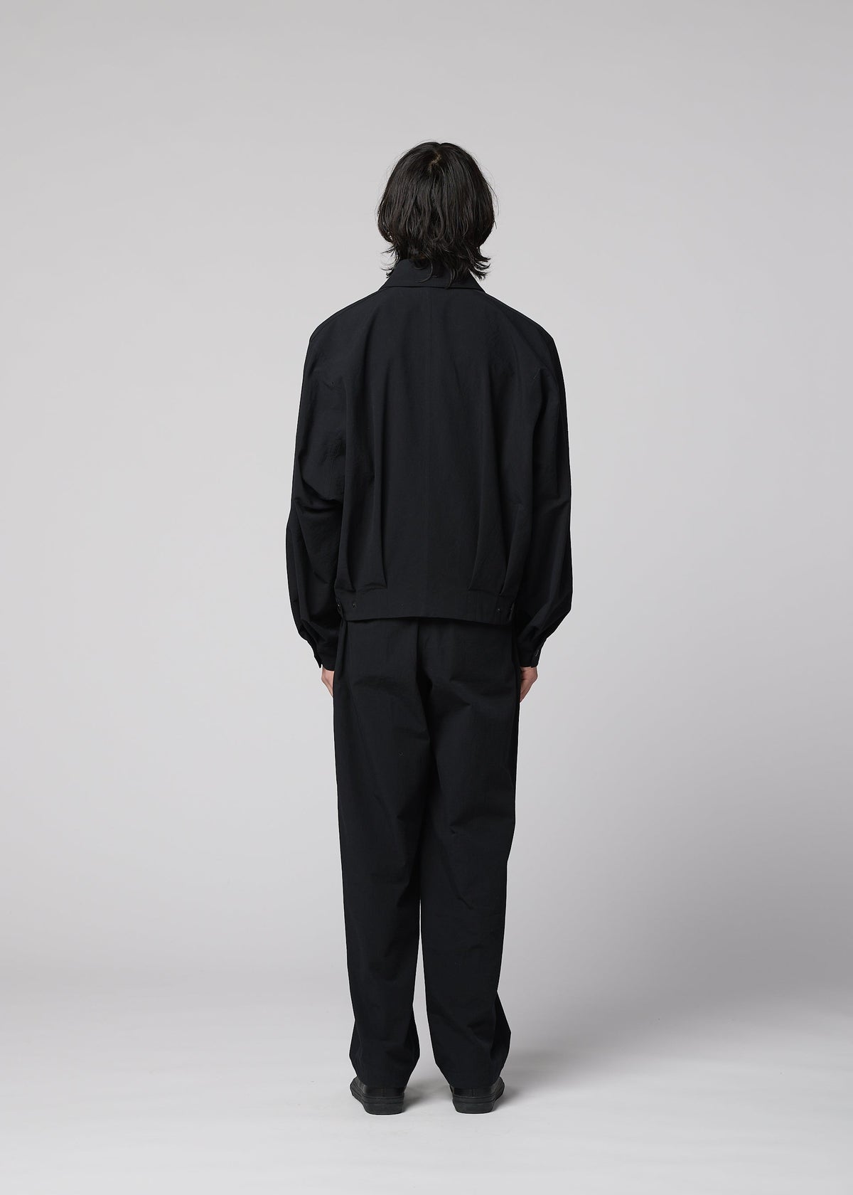 A person stands facing away from the camera, wearing IM MEN’s COMFORT COTTON loose-fitting black jacket and high-twist organic cotton straight pants, which offer a smooth feel against a plain light gray background. view 7 of 8