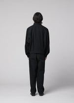 A person stands facing away from the camera, wearing IM MEN’s COMFORT COTTON loose-fitting black jacket and high-twist organic cotton straight pants, which offer a smooth feel against a plain light gray background.