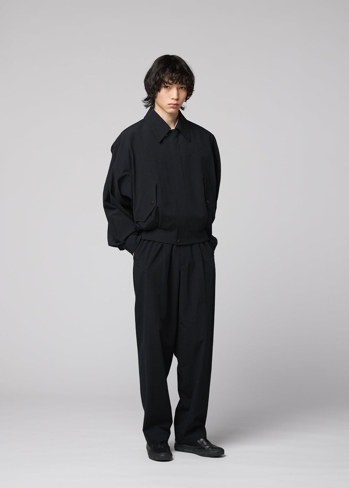 A person with shaggy hair stands against a plain light gray background, wearing the IM MEN COMFORT COTTON loose-fitting black jacket and matching wide straight pants crafted from high-twist organic cotton for a smooth feel, paired with black shoes and their hands in their pockets. view 8 of 8