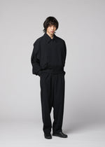 A person with shaggy hair stands against a plain light gray background, wearing the IM MEN COMFORT COTTON loose-fitting black jacket and matching wide straight pants crafted from high-twist organic cotton for a smooth feel, paired with black shoes and their hands in their pockets.