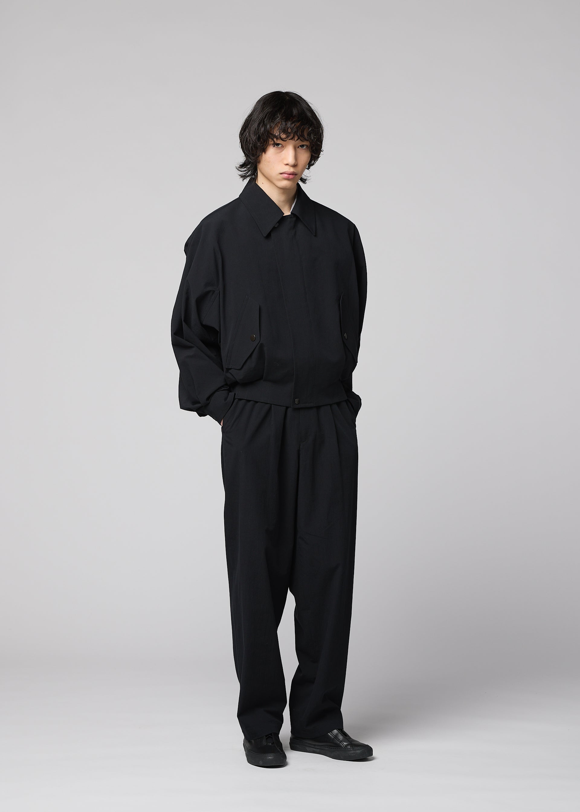 Zoomed view A person with shaggy hair stands against a plain light gray background, wearing the IM MEN COMFORT COTTON loose-fitting black jacket and matching wide straight pants crafted from high-twist organic cotton for a smooth feel, paired with black shoes and their hands in their pockets.