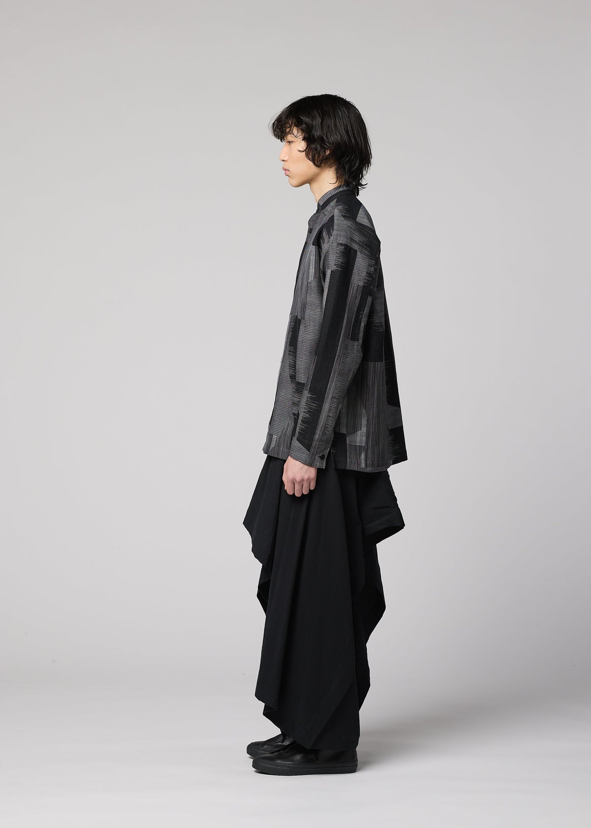 A person with medium-length dark hair stands in profile against a plain light gray background, wearing IM MEN’s COMFORT COTTON: a gray patterned jacket paired with long, draped irregular-cut pants evocative of a skirt, both crafted from high-twist organic cotton. view 6 of 8