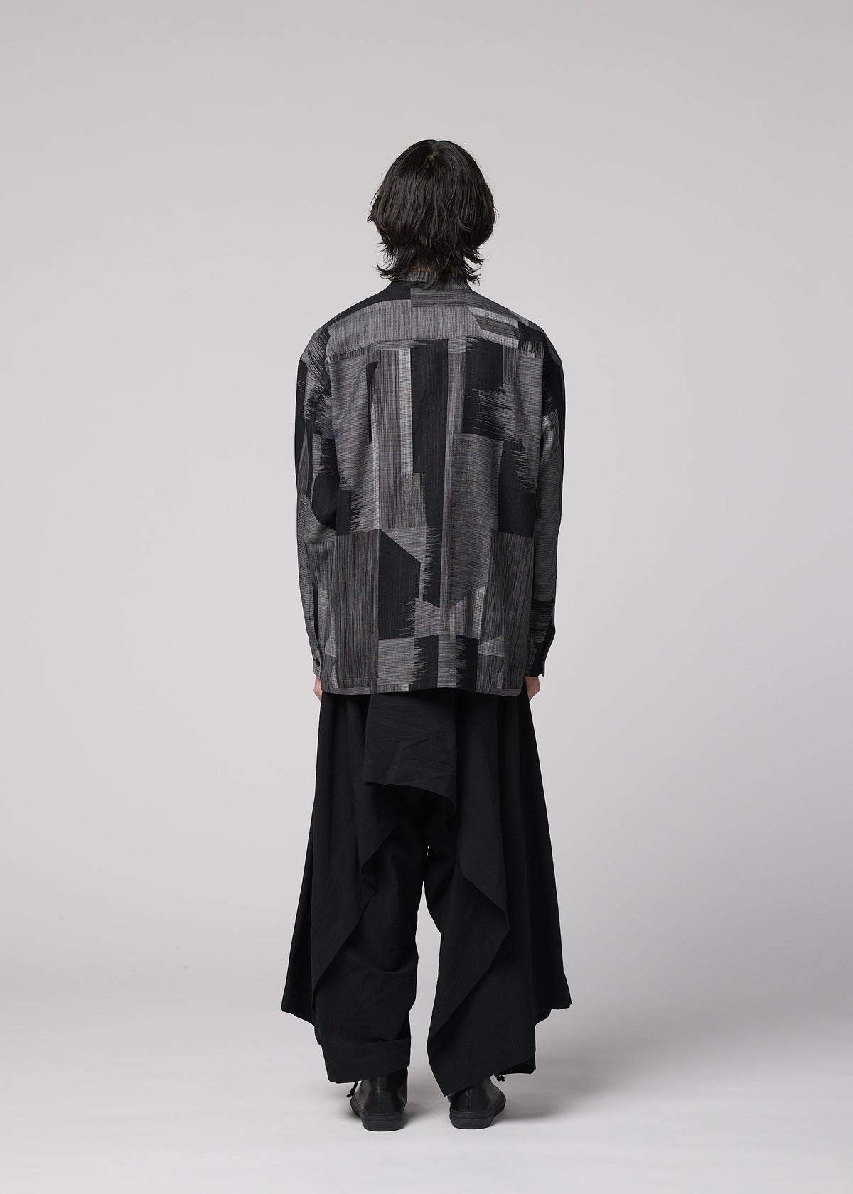 A person with medium-length dark hair stands facing away, wearing IM MEN’s COMFORT COTTON patterned gray and black jacket crafted from high-twist organic cotton, paired with loose irregular-cut black pants, against a plain light gray background. view 7 of 8