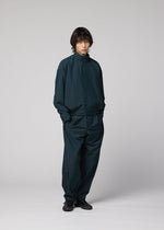 A person with medium-length dark hair stands against a light grey background, modeling IM MEN's FLUENT: a loose-fitting dark teal jacket and wrinkle-resistant pants, paired with black shoes, hands in pockets and looking at the camera.