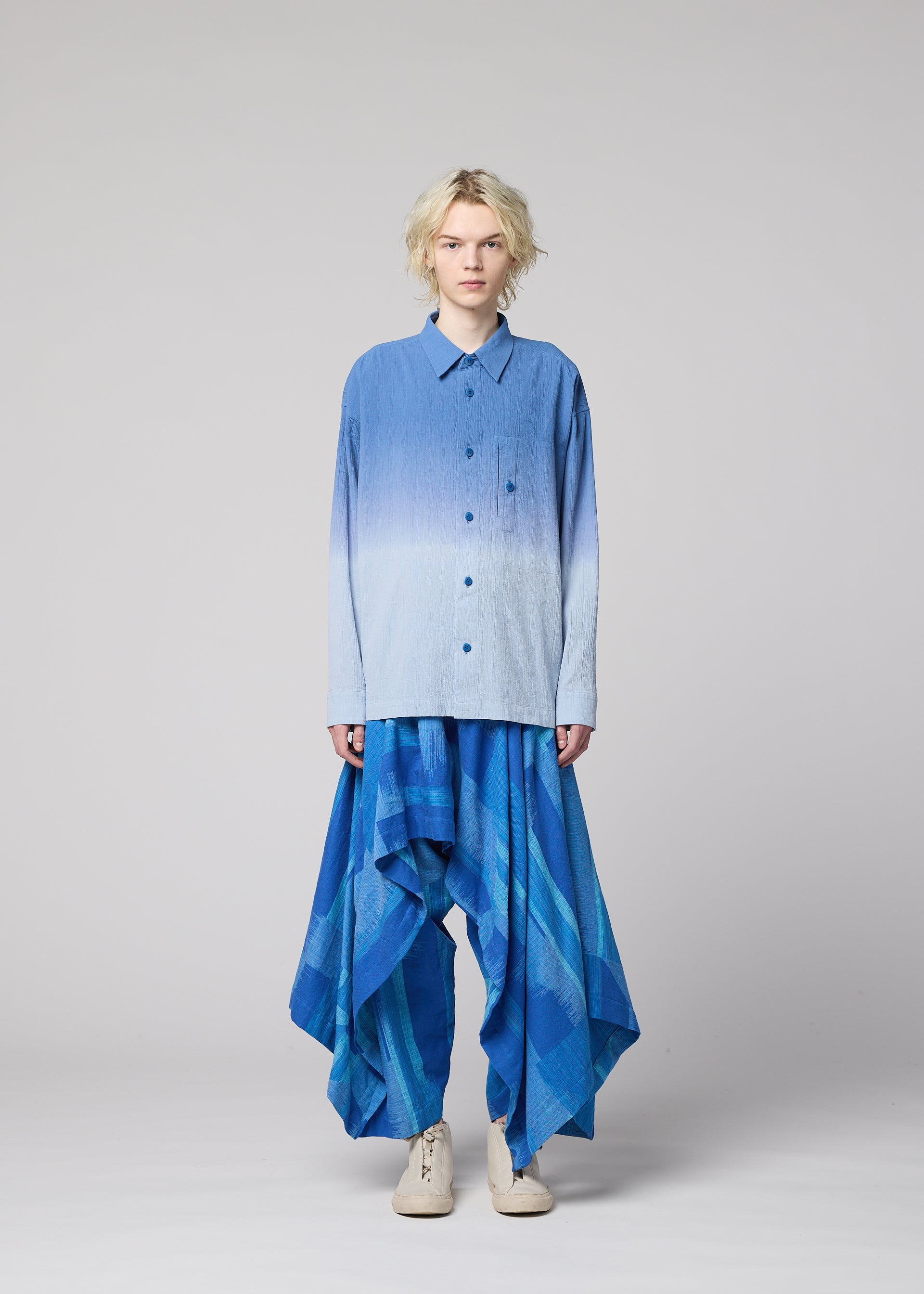 Zoomed view A person wearing the IM MEN KASURI loose gradient blue button-up shirt and wide, flowing pants—crafted with traditional Kasuri weaving—stands against a plain light gray background.