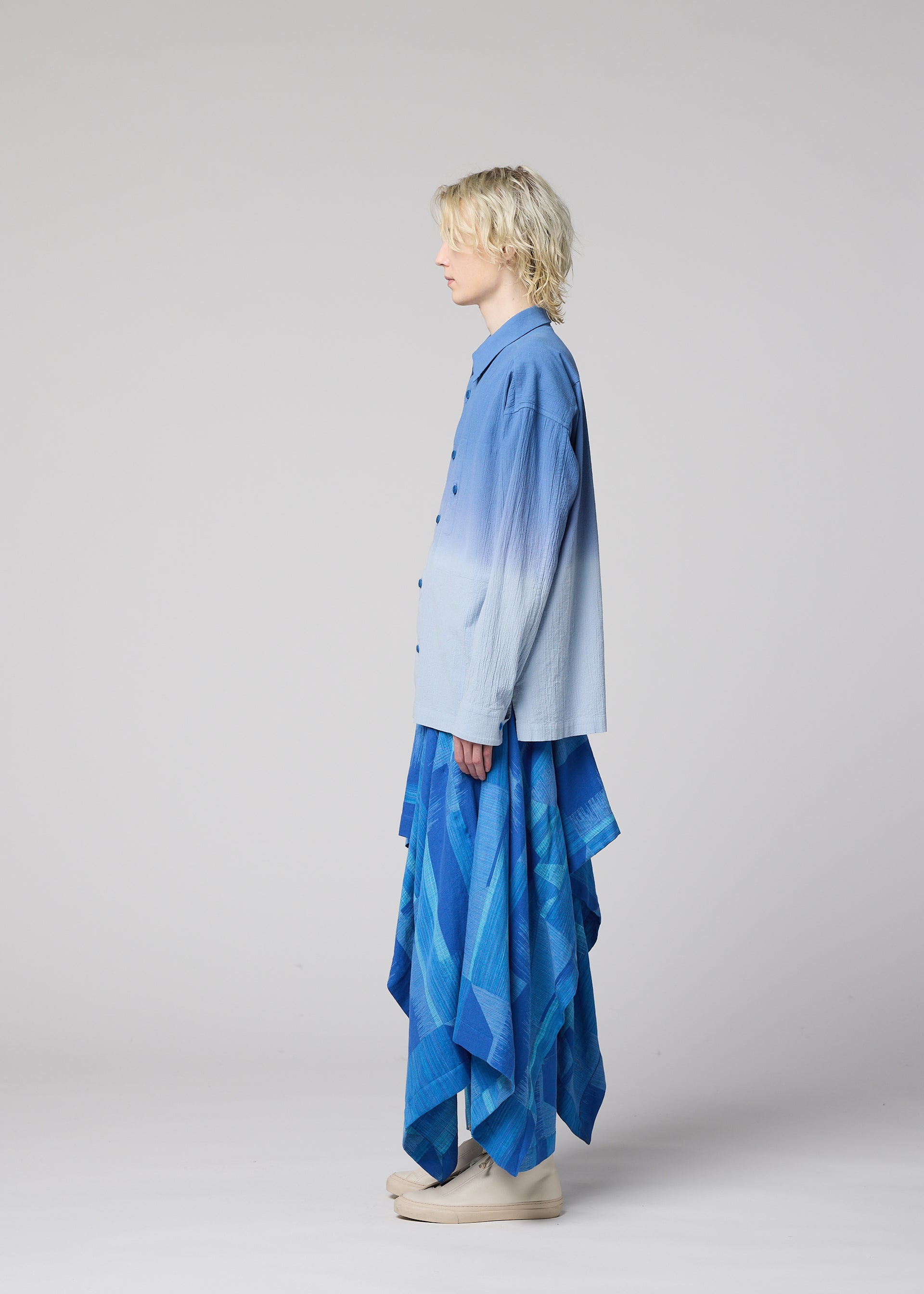 Zoomed view A person with blond hair stands in profile, dressed in the IM MEN KASURI blue button-up shirt and layered skirt featuring geometric patterns inspired by Kasuri weaving, paired with light-colored shoes, against a plain light background.
