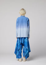 A person with short, curly blonde hair stands facing away on a plain light gray background, wearing IM MEN’s layered, asymmetric KASURI pants—design inspired by traditional Kasuri weaving—paired with a blue ombre shirt and white shoes.