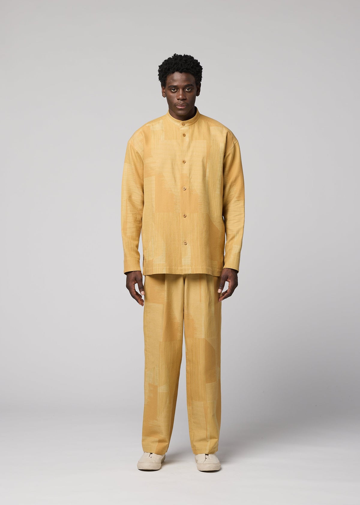 A man with short curly hair stands facing forward against a plain gray background, dressed in the IM MEN KASURI set—a loose-fitting, light yellow button-up shirt and matching pants featuring a subtle pattern inspired by traditional Kasuri weaving. He completes the look with off-white shoes. view 4 of 7