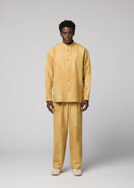 A man with short curly hair stands facing forward against a plain gray background, dressed in the IM MEN KASURI set—a loose-fitting, light yellow button-up shirt and matching pants featuring a subtle pattern inspired by traditional Kasuri weaving. He completes the look with off-white shoes.