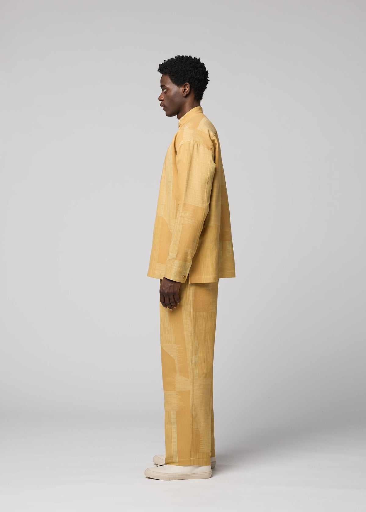 A person stands in profile against a plain background, wearing the IM MEN KASURI set: a loose-fitting, light yellow patterned long-sleeve top paired with matching Kasuri pants. Both pieces display the distinctive textures of traditional Kasuri weaving, completed with light shoes. view 5 of 7