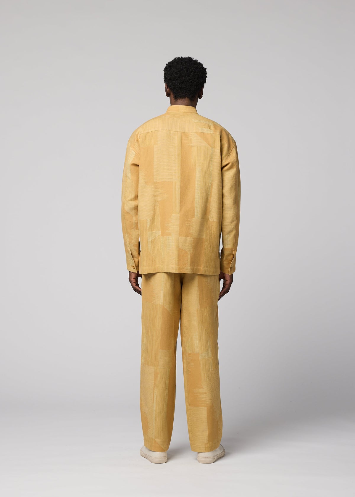 A person stands facing away, wearing a loose-fitting, long-sleeved, light yellow patterned IM MEN KASURI shirt paired with matching KASURI pants and beige shoes. The background is light gray and minimalistic. view 6 of 7