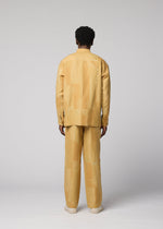 A person stands facing away, wearing a loose-fitting, long-sleeved, light yellow patterned IM MEN KASURI shirt paired with matching KASURI pants and beige shoes. The background is light gray and minimalistic.