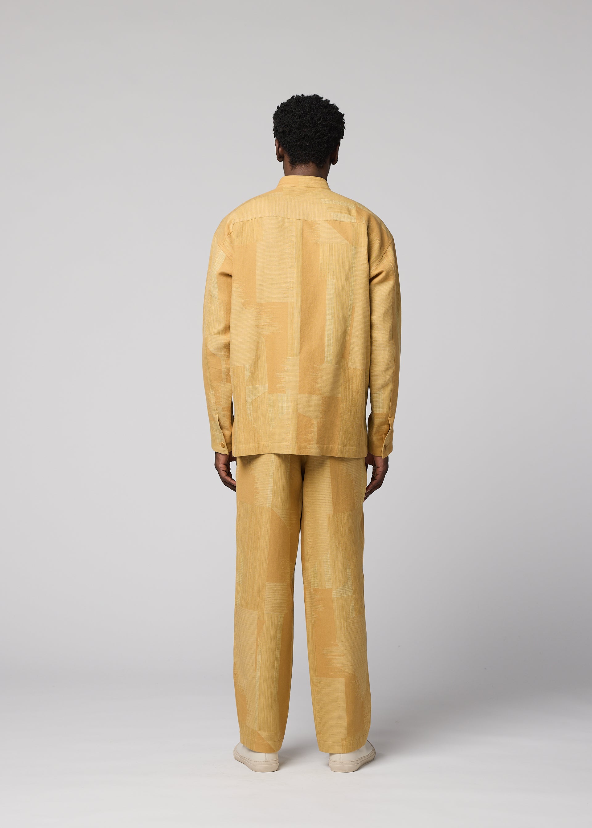Zoomed view A person stands facing away, wearing a loose-fitting, long-sleeved, light yellow patterned IM MEN KASURI shirt paired with matching KASURI pants and beige shoes. The background is light gray and minimalistic.