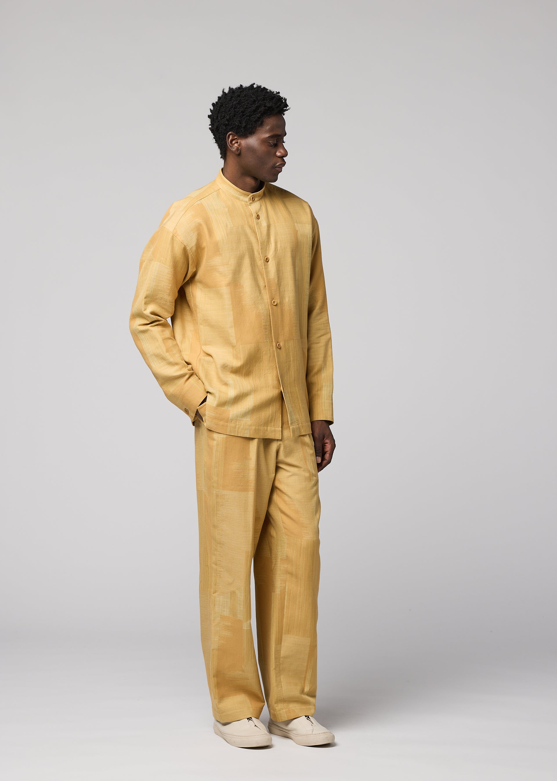 Zoomed view A man stands sideways against a plain light gray background, wearing IM MEN’s KASURI set—a matching light gold button-up shirt and pants, crafted with the intricate beauty of Kasuri weaving—paired with white shoes.