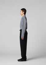 A person with short hair stands in profile against a plain light background, wearing a gray button-up shirt, IM MEN SHRINK WOOL black tapered pants, and black shoes, arms relaxed at their sides.