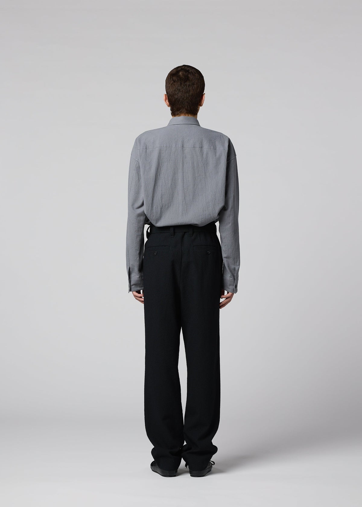 A person faces away from the camera, wearing a gray long-sleeve shirt and black SHRINK WOOL pants by IM MEN, against a plain light gray background. view 8 of 9