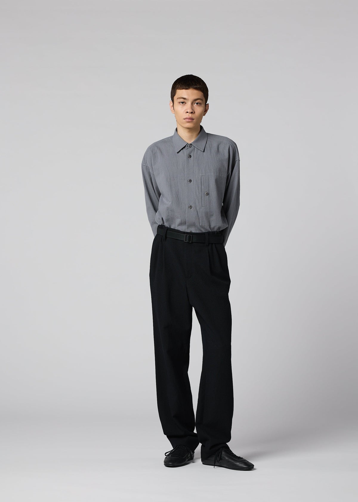A person stands against a plain light gray background, wearing a loose-fitting gray button-up shirt, black SHRINK WOOL pants by IM MEN, and black shoes, with their hands behind their back. view 9 of 9