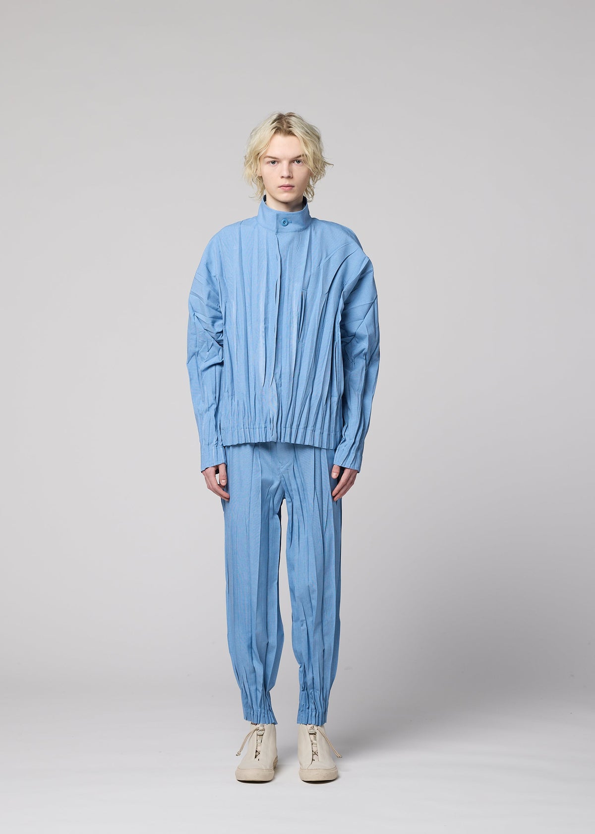 A person with wavy blonde hair stands facing forward, wearing the EDGE pleated jacket and matching hand-gathered pleated pants in light blue mélange polyester from IM MEN, paired with white shoes, against a plain light gray background. view 6 of 9
