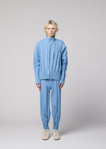 A person with wavy blonde hair stands facing forward, wearing the EDGE pleated jacket and matching hand-gathered pleated pants in light blue mélange polyester from IM MEN, paired with white shoes, against a plain light gray background.