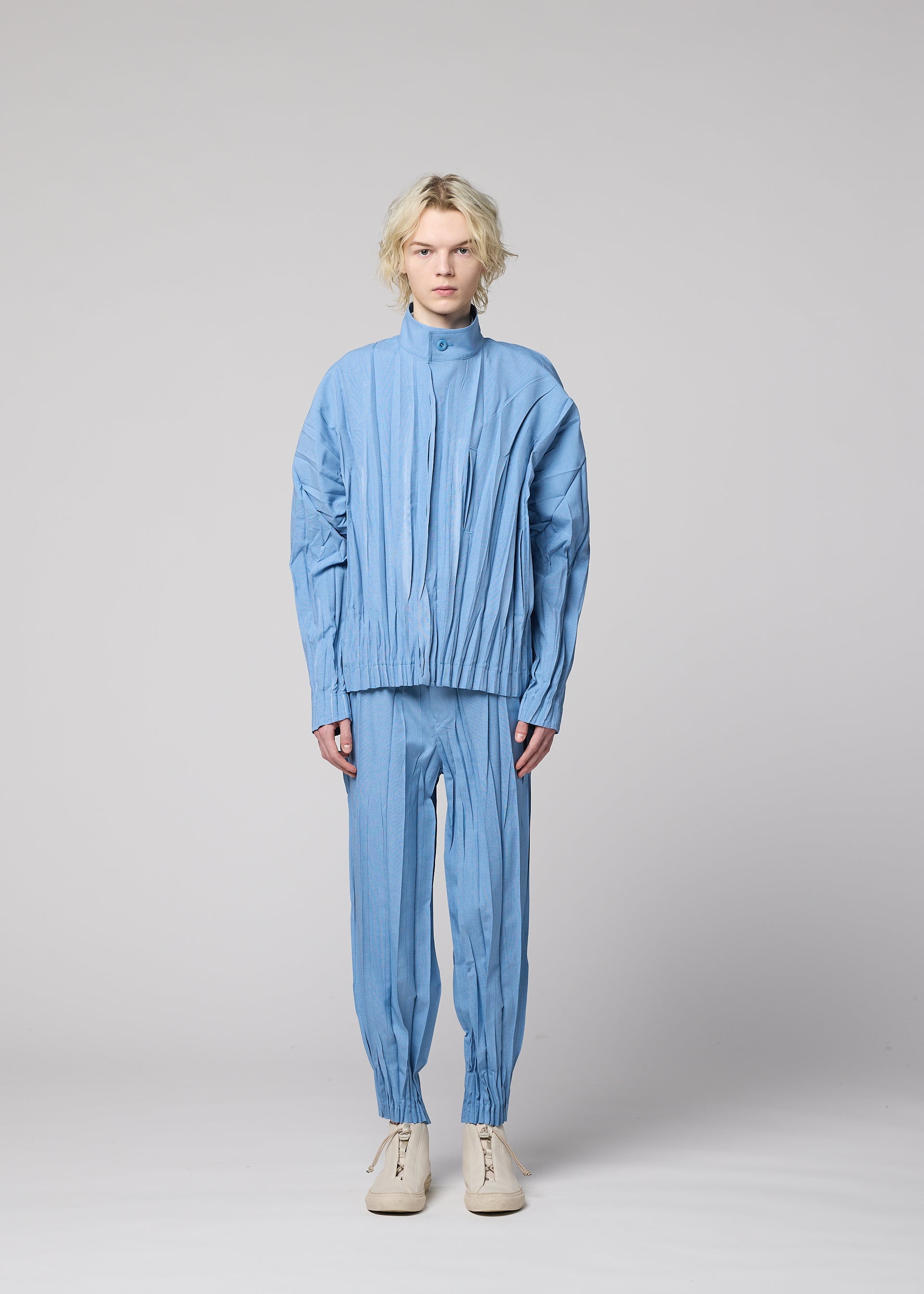 Zoomed view A person with wavy blonde hair stands facing forward, wearing the EDGE pleated jacket and matching hand-gathered pleated pants in light blue mélange polyester from IM MEN, paired with white shoes, against a plain light gray background.