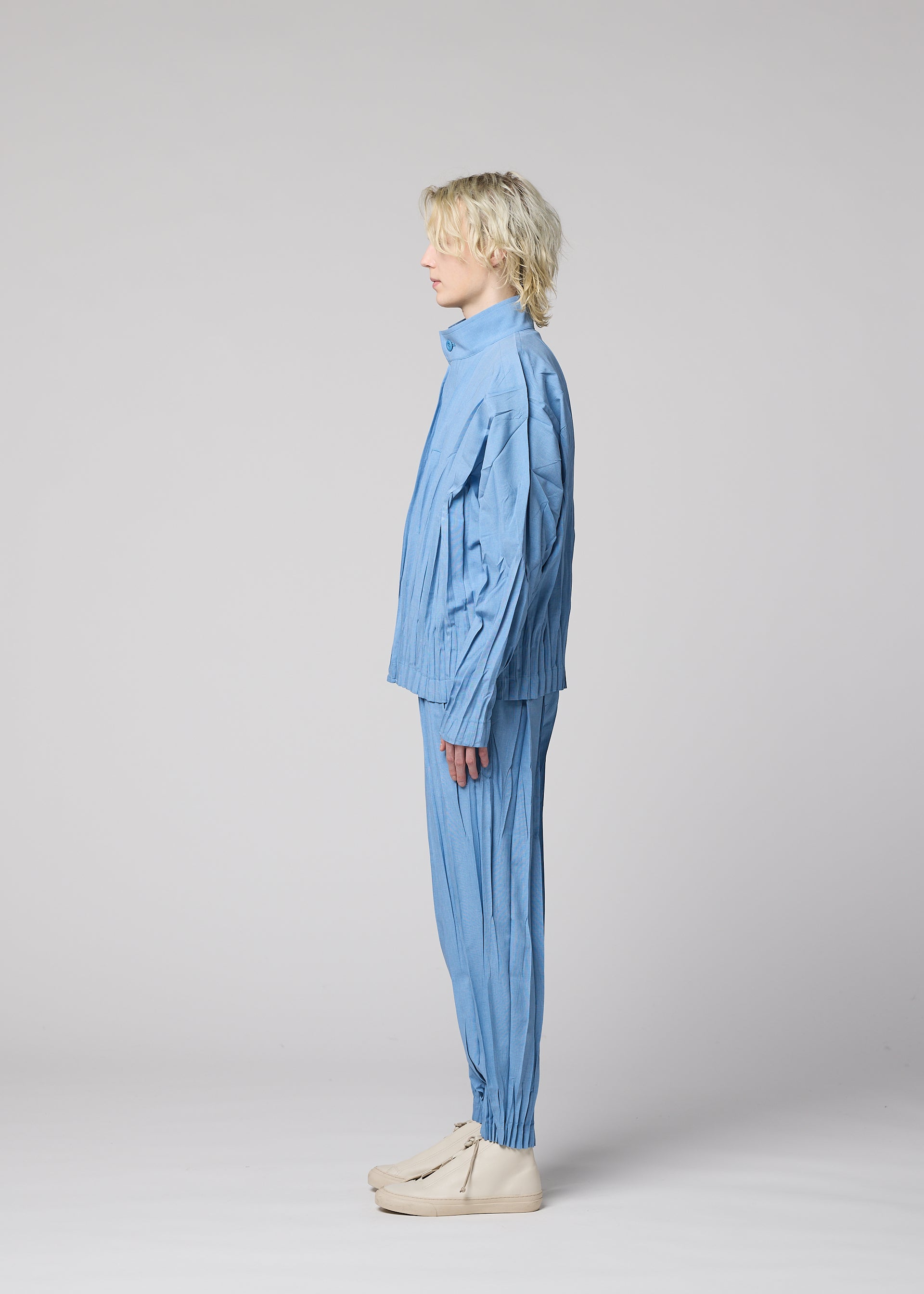 Zoomed view A person with light wavy hair stands in profile against a plain light gray background, wearing IM MEN’s EDGE—a loose-fitting blue pleated ensemble made from hand-gathered mélange polyester pants—paired with cream shoes.