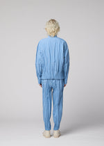 A person with short, wavy blonde hair stands facing away, wearing the EDGE light blue crinkled mélange polyester tracksuit from IM MEN and cream shoes, set against a plain light gray background.