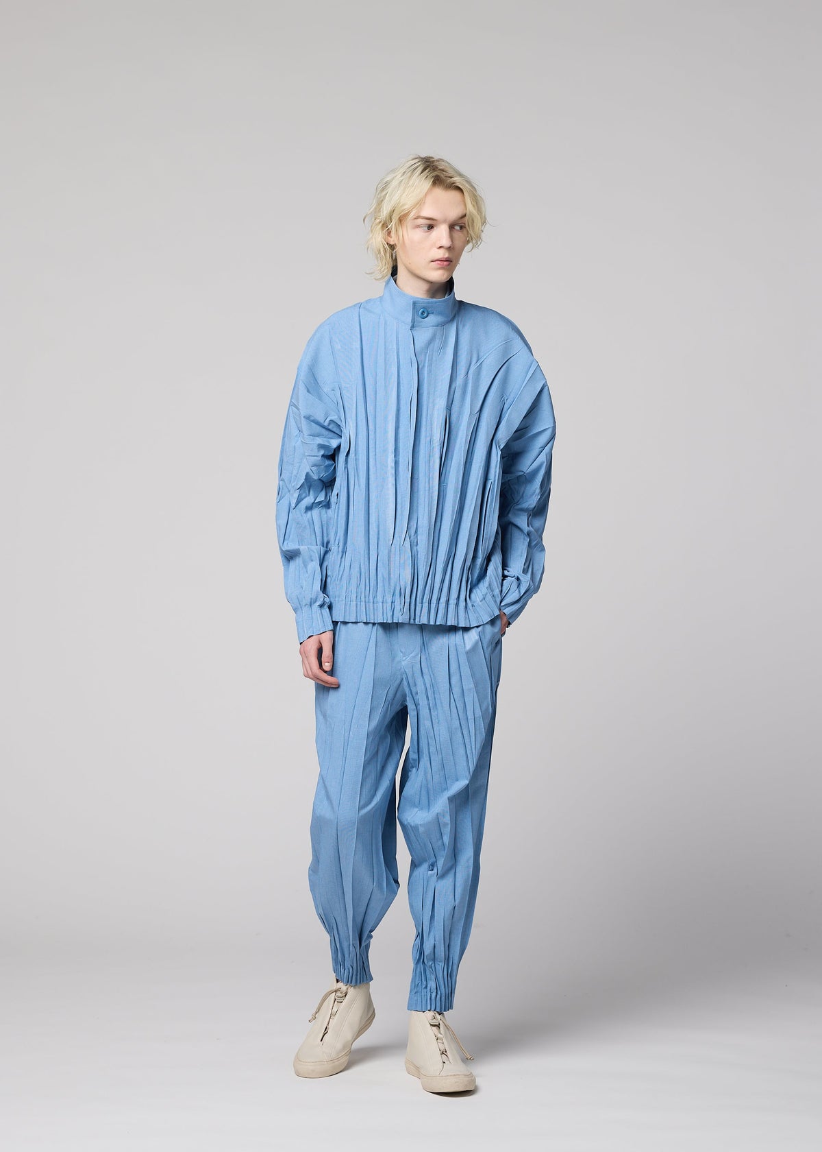 A person with blond hair wears the EDGE light blue pleated jacket and hand-gathered pleated pants in mélange polyester by IM MEN, paired with white shoes, standing against a plain light gray background. view 9 of 9