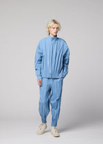 A person with blond hair wears the EDGE light blue pleated jacket and hand-gathered pleated pants in mélange polyester by IM MEN, paired with white shoes, standing against a plain light gray background.