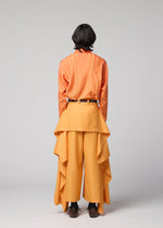 A person with shoulder-length dark hair stands facing away against a plain light gray background, wearing the IM MEN BREEZE DRAPE wide-legged flowy pants with draped fabric, paired with an orange shirt with yellow stripes.