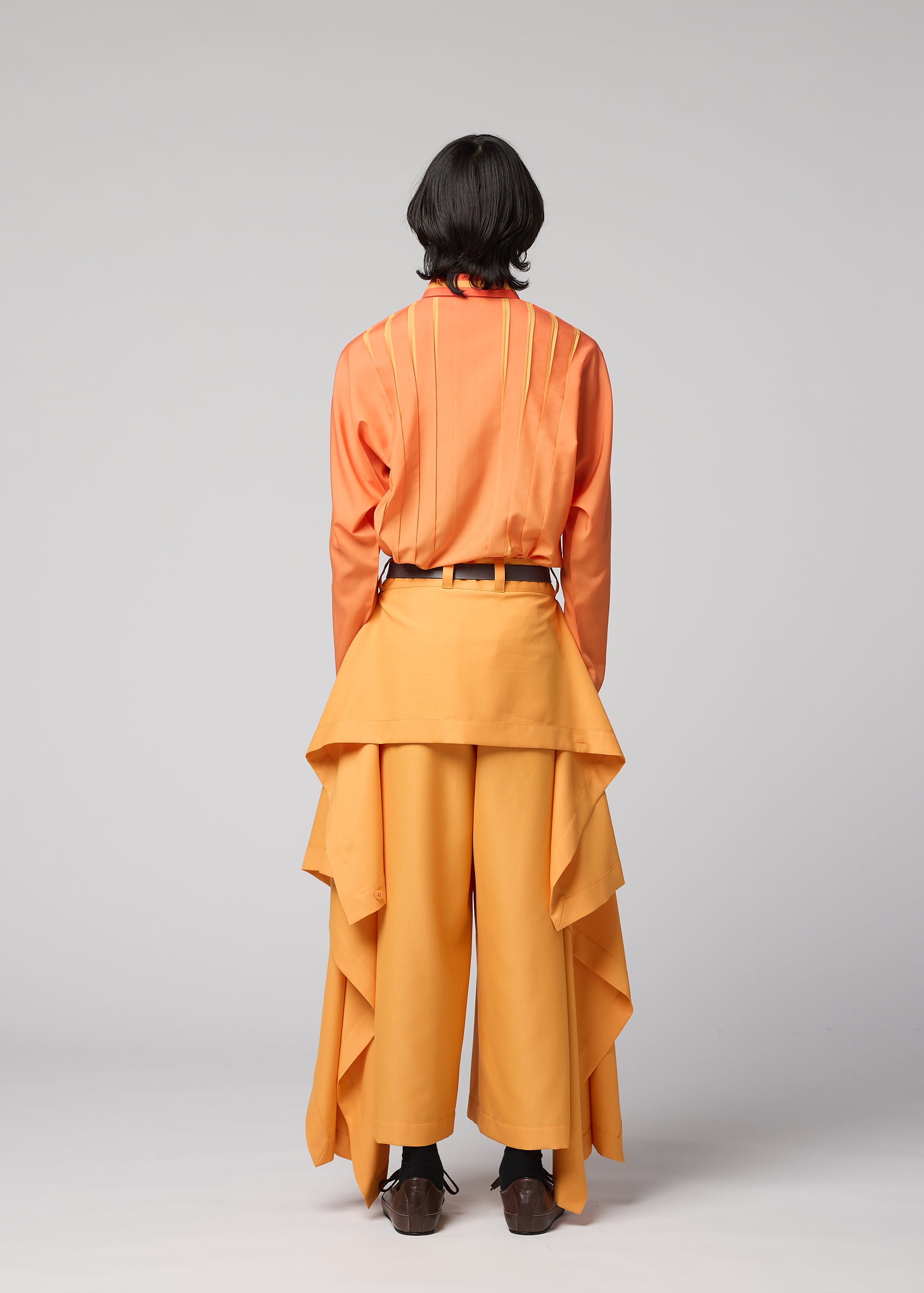 Zoomed view A person with shoulder-length dark hair stands facing away against a plain light gray background, wearing the IM MEN BREEZE DRAPE wide-legged flowy pants with draped fabric, paired with an orange shirt with yellow stripes.