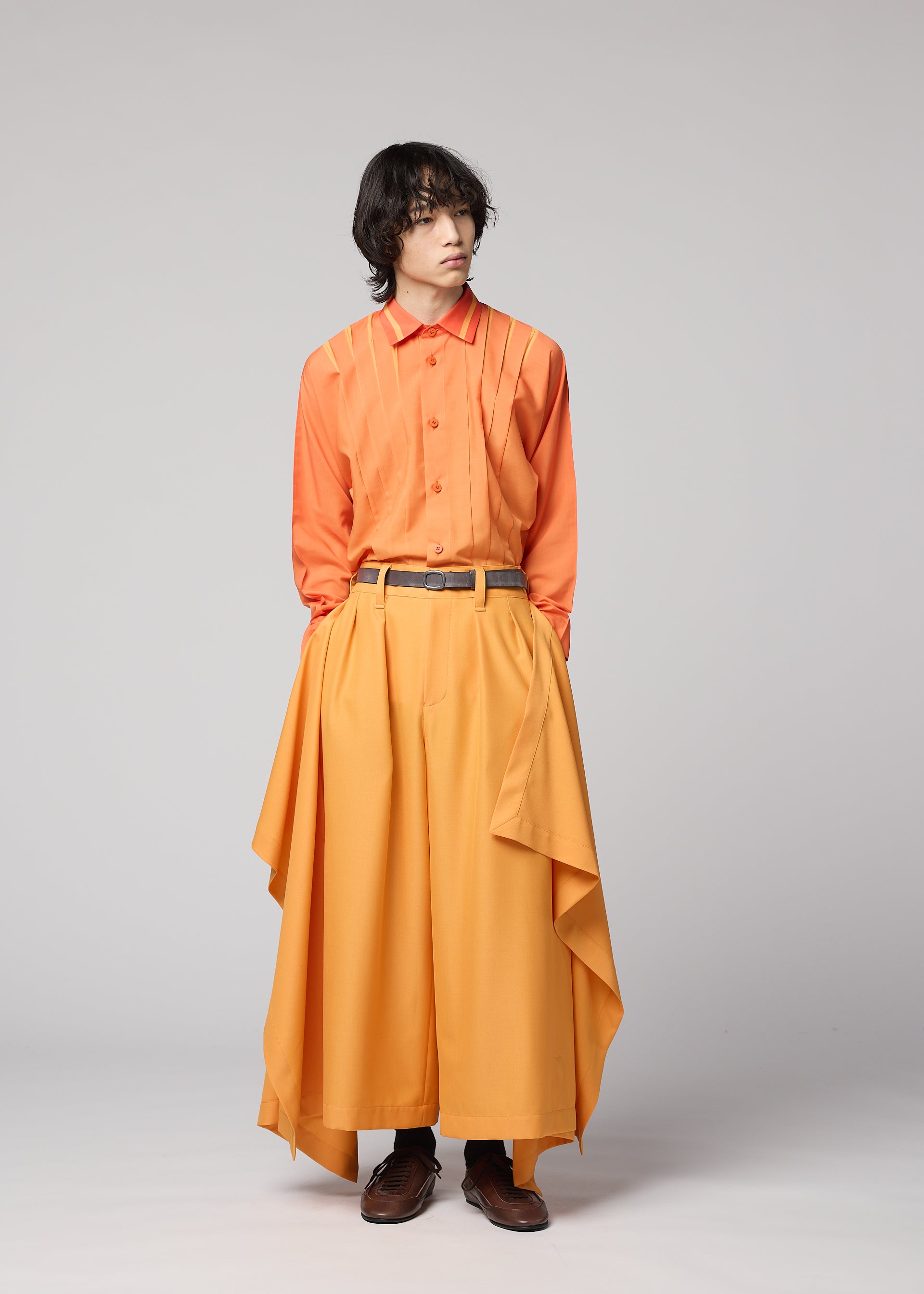 Zoomed view A person with medium-length dark hair wears an orange button-up shirt and the IM MEN BREEZE DRAPE pants, featuring beautifully layered, wide, and flowy fabric. They stand against a plain gray background, looking to the side with hands in their pockets.