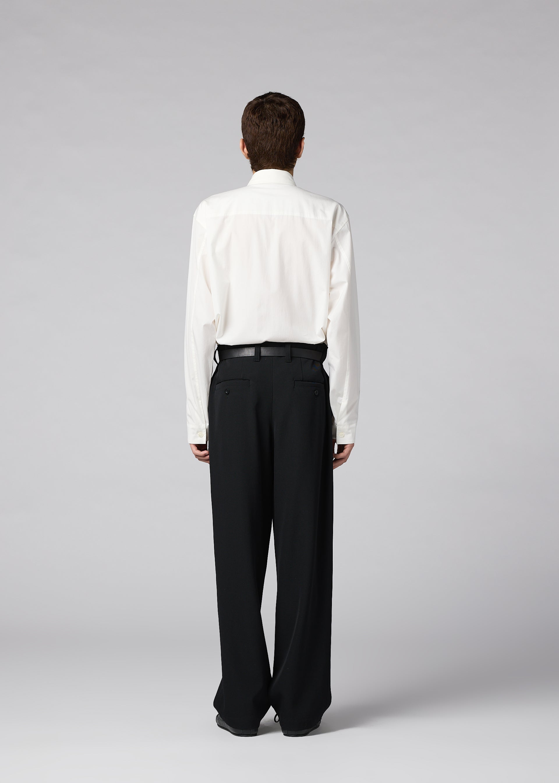 Zoomed view A person with short brown hair stands facing away, wearing IM MEN’s FRONT BACK white dress shirt tucked into matte wool trousers with a black belt, against a plain light gray background.