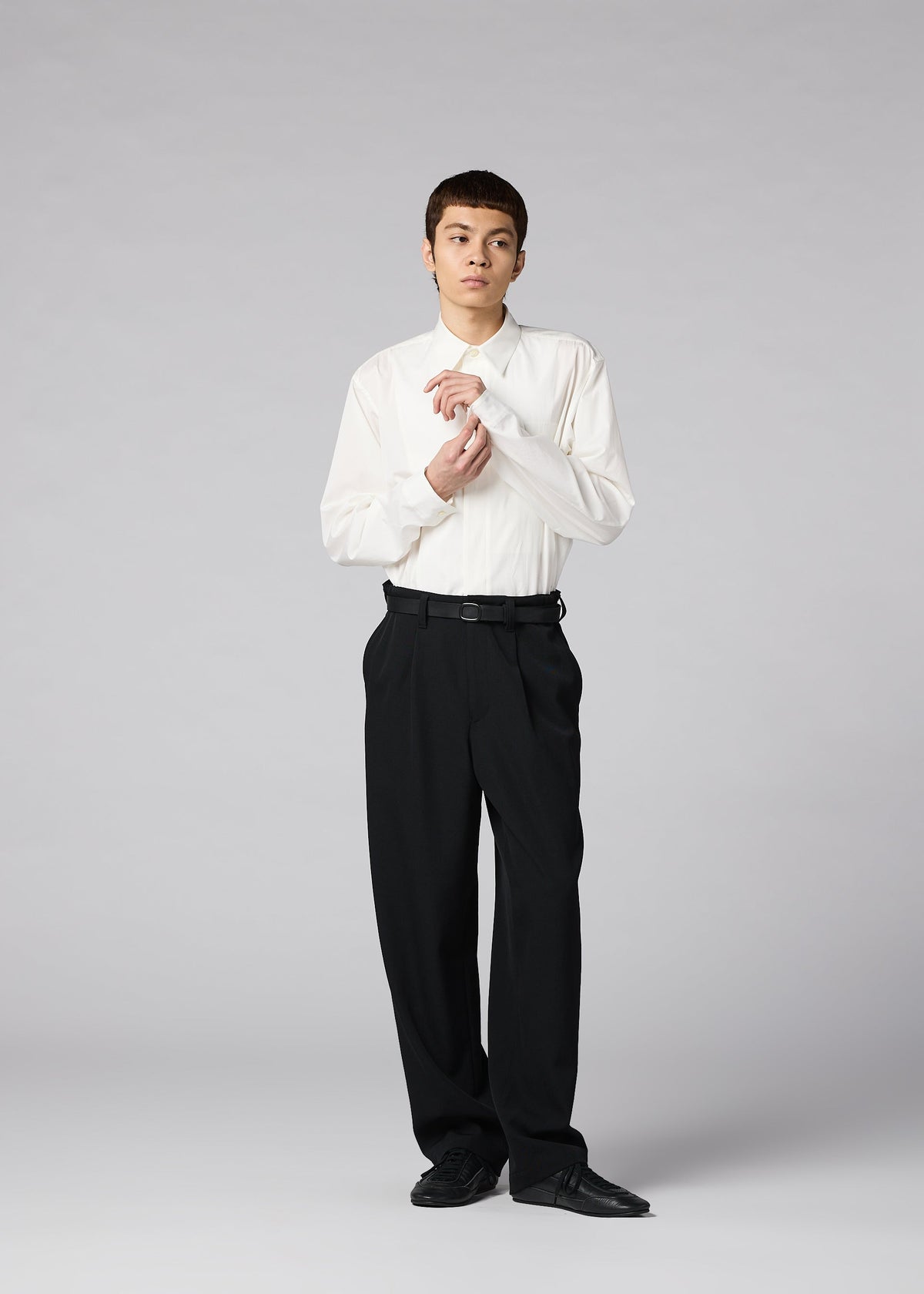 A person stands against a gray background wearing the IM MEN FRONT BACK white dress shirt, matte wool trousers, and black shoes, adjusting their cuff with a neutral expression. view 7 of 7