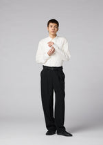 A person stands against a gray background wearing the IM MEN FRONT BACK white dress shirt, matte wool trousers, and black shoes, adjusting their cuff with a neutral expression.