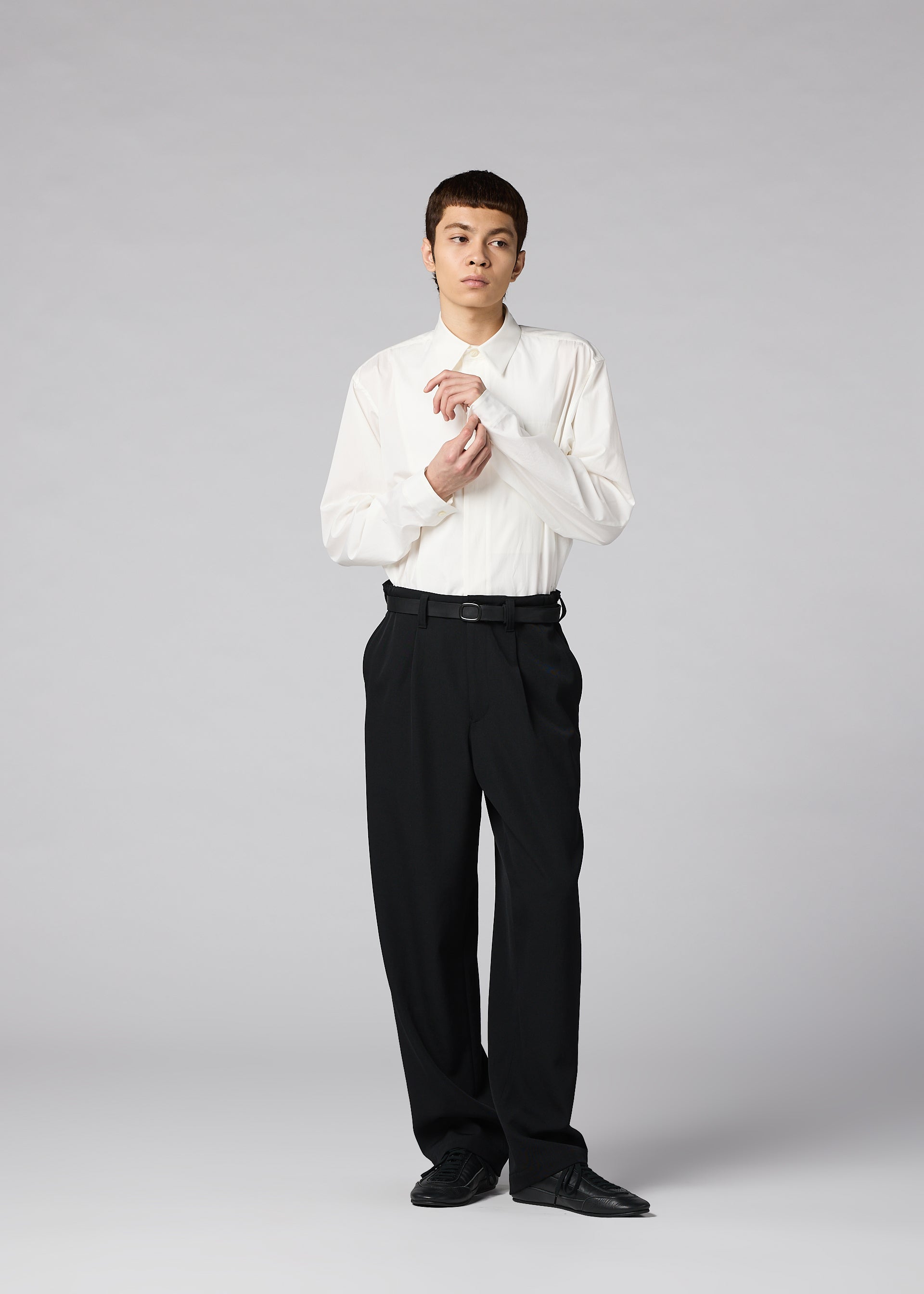 Zoomed view A person stands against a gray background wearing the IM MEN FRONT BACK white dress shirt, matte wool trousers, and black shoes, adjusting their cuff with a neutral expression.