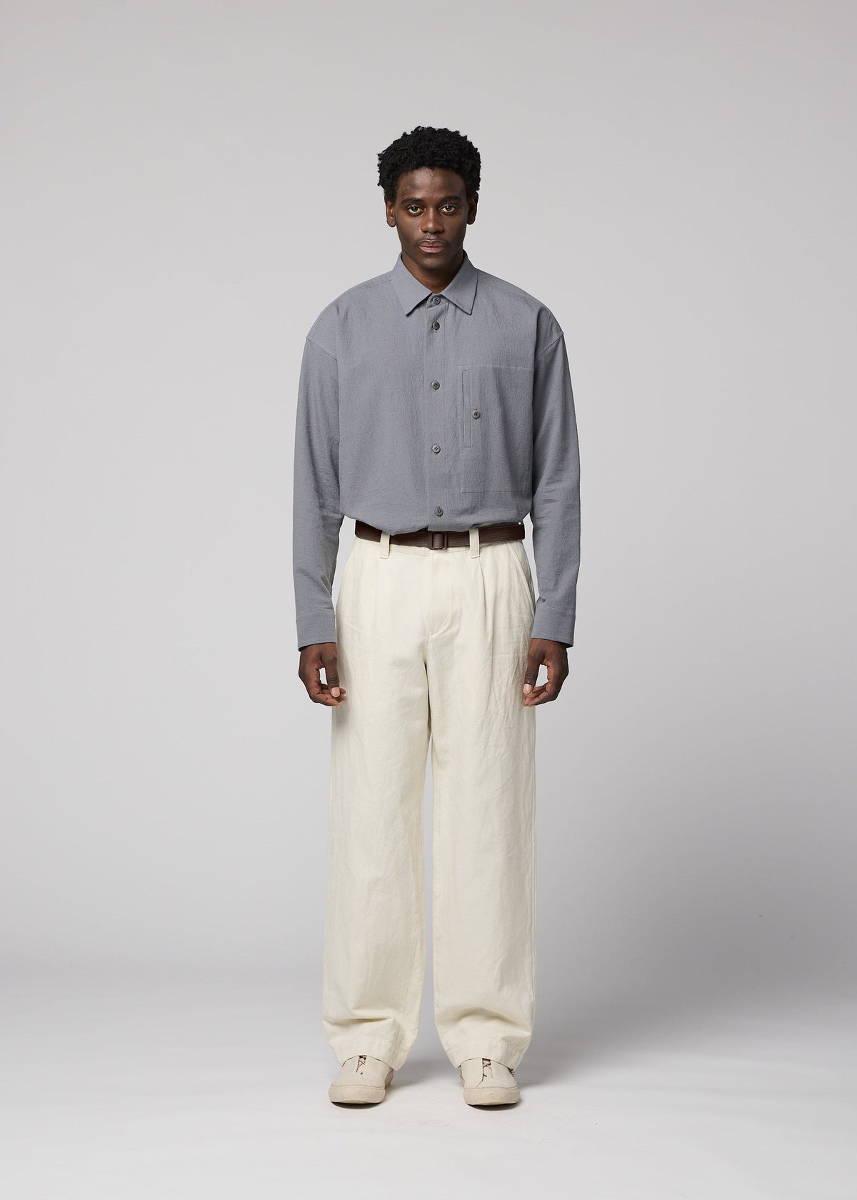 A man stands facing forward against a plain light gray background, wearing an IM MEN TOUGH WOOL COTTON gray button-up shirt tucked into high-waisted, loose-fitting cream Bishu wool textiles trousers, styled with a brown belt and beige shoes. view 6 of 9