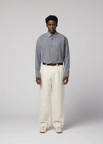 A man stands facing forward against a plain light gray background, wearing an IM MEN TOUGH WOOL COTTON gray button-up shirt tucked into high-waisted, loose-fitting cream Bishu wool textiles trousers, styled with a brown belt and beige shoes.