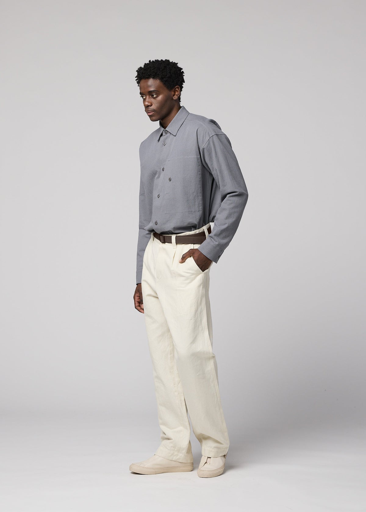 A man stands against a plain light background, wearing a gray button-up shirt tucked into cream-colored IM MEN TOUGH WOOL COTTON trousers with a belt. His left hand is in his pocket, and he is looking slightly to the left. view 9 of 9