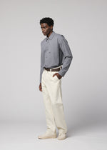 A man stands against a plain light background, wearing a gray button-up shirt tucked into cream-colored IM MEN TOUGH WOOL COTTON trousers with a belt. His left hand is in his pocket, and he is looking slightly to the left.