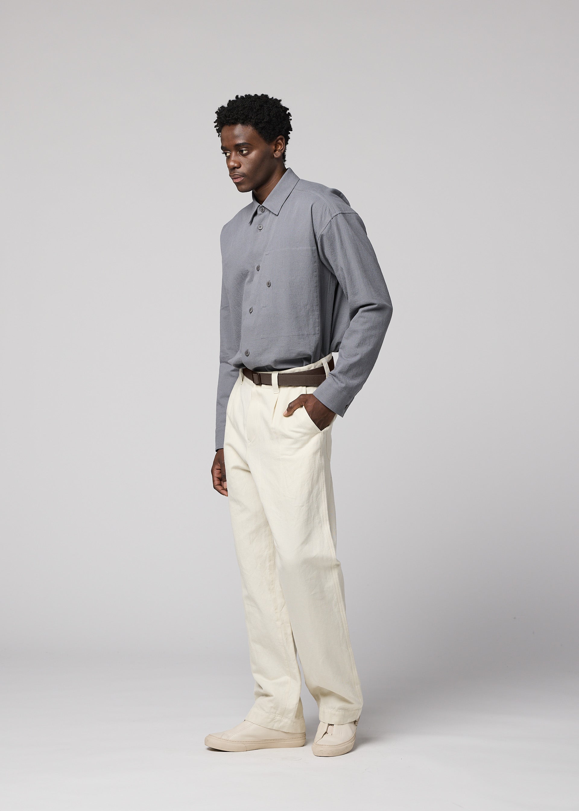 Zoomed view A man stands against a plain light background, wearing a gray button-up shirt tucked into cream-colored IM MEN TOUGH WOOL COTTON trousers with a belt. His left hand is in his pocket, and he is looking slightly to the left.