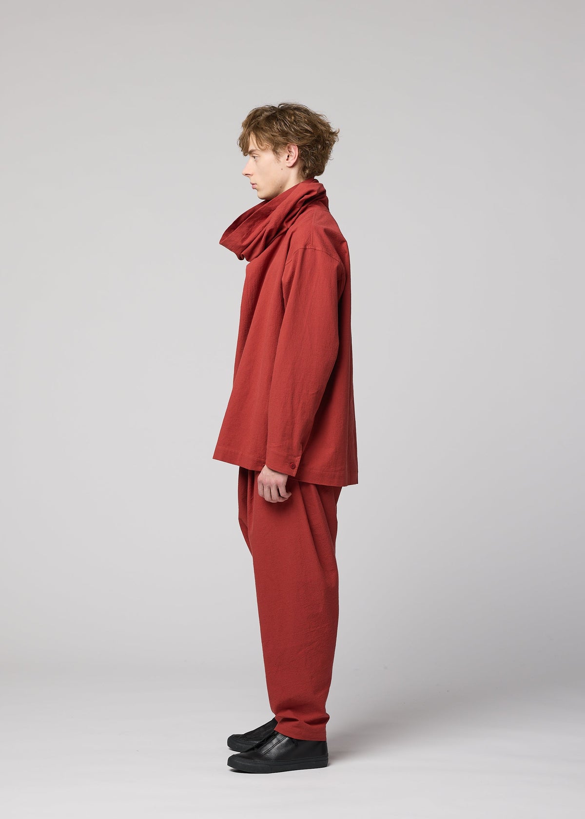 A person stands in profile against a plain gray background, dressed in loose-fitting, matching red clothing with a relaxed silhouette and high collar, paired with black shoes. The look highlights the COTTON YAK pants from IM MEN. view 7 of 9