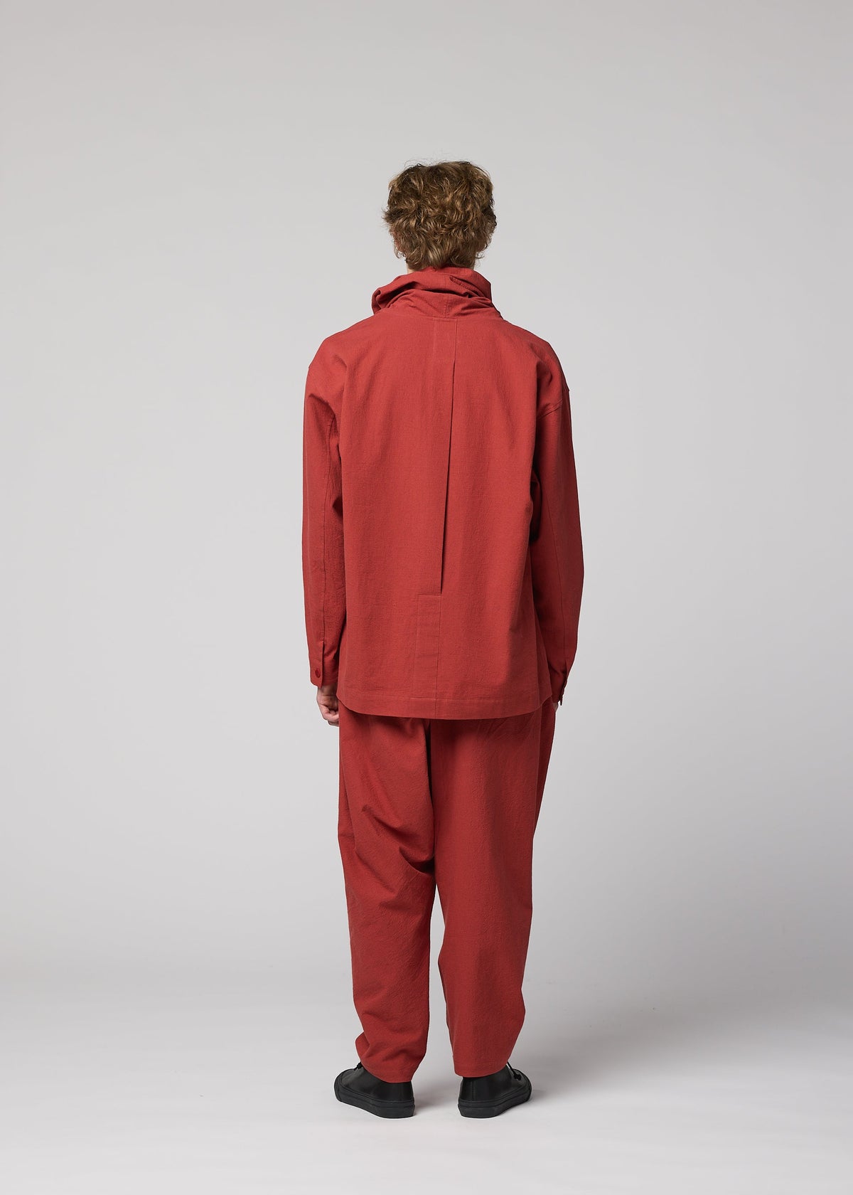 A person with curly hair faces away, dressed in a loose-fitting red COTTON YAK jacket and matching pants from IM MEN, creating a relaxed silhouette paired with black shoes against a plain light gray background. This look is featured in the IM MEN Autumn Winter 2026/27 Collection. view 8 of 9