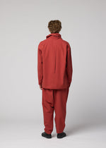 A person with curly hair faces away, dressed in a loose-fitting red COTTON YAK jacket and matching pants from IM MEN, creating a relaxed silhouette paired with black shoes against a plain light gray background. This look is featured in the IM MEN Autumn Winter 2026/27 Collection.