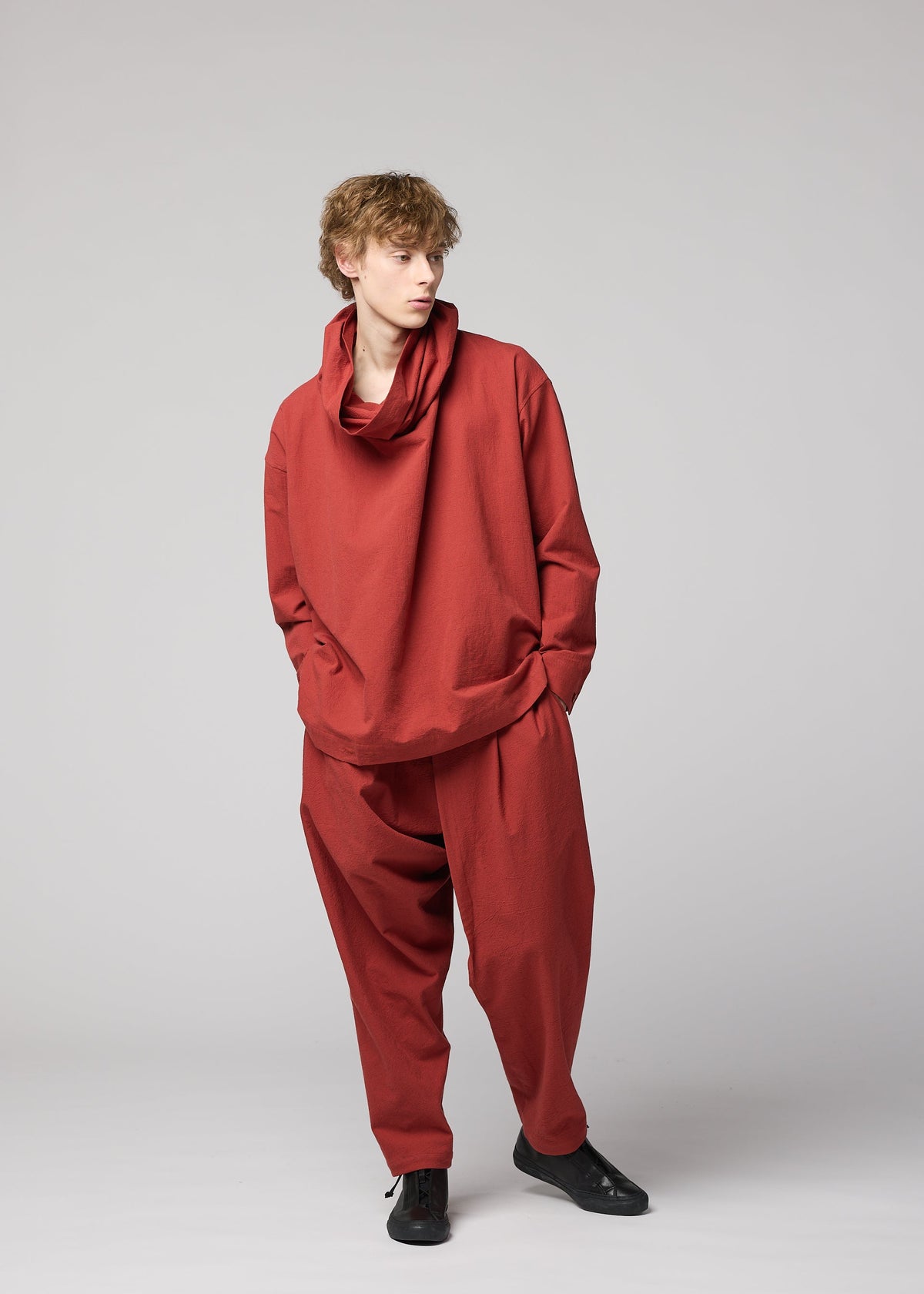 Wearing the IM MEN COTTON YAK set from the Autumn Winter 2026/27 Collection, this person showcases a relaxed silhouette in a loose-fitting, long-sleeved red top with a draped collar and matching baggy cotton yak pants. Standing against a plain light gray background with hands in pockets, they look to the left. view 9 of 9