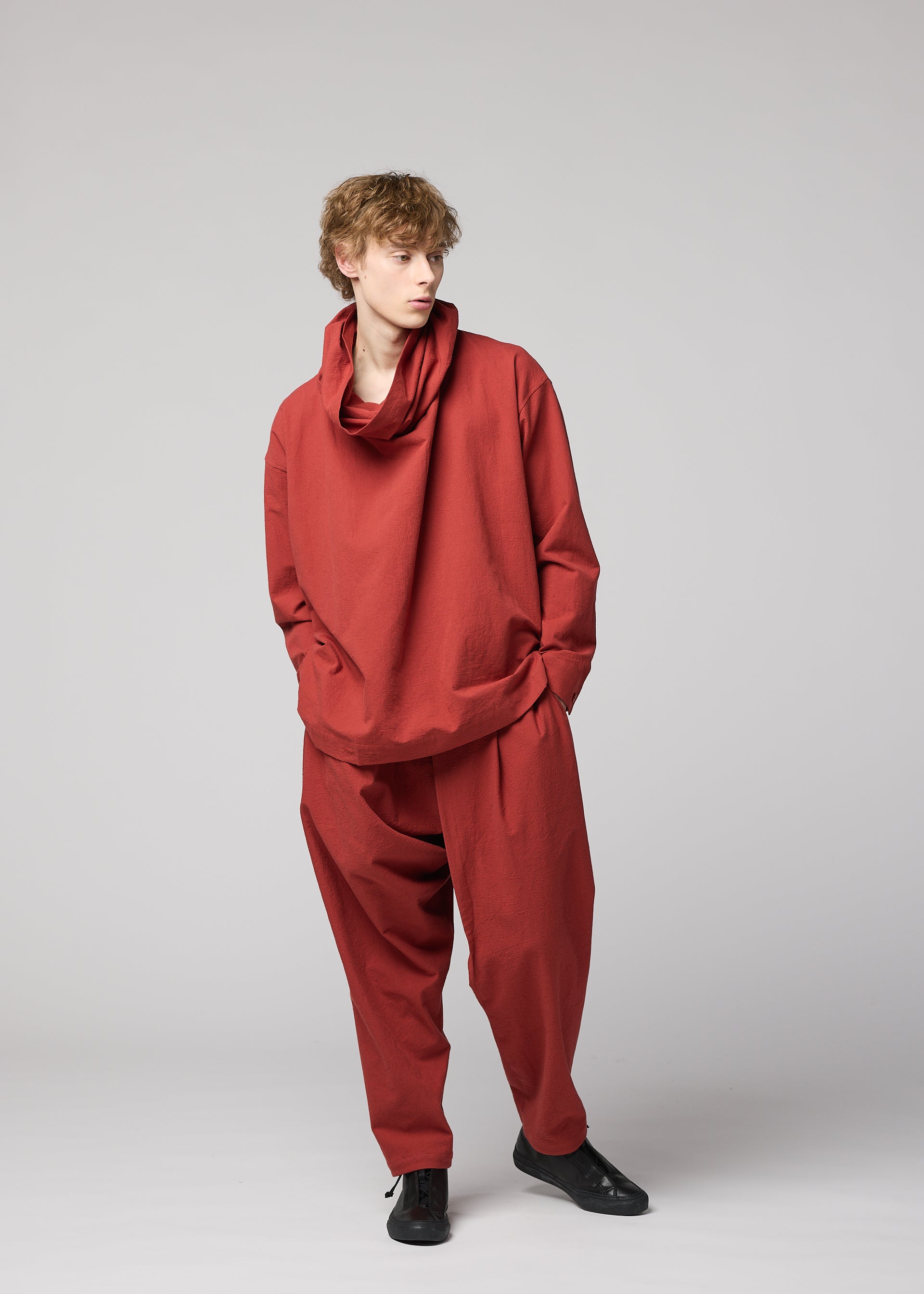 Zoomed view Wearing the IM MEN COTTON YAK set from the Autumn Winter 2026/27 Collection, this person showcases a relaxed silhouette in a loose-fitting, long-sleeved red top with a draped collar and matching baggy cotton yak pants. Standing against a plain light gray background with hands in pockets, they look to the left.
