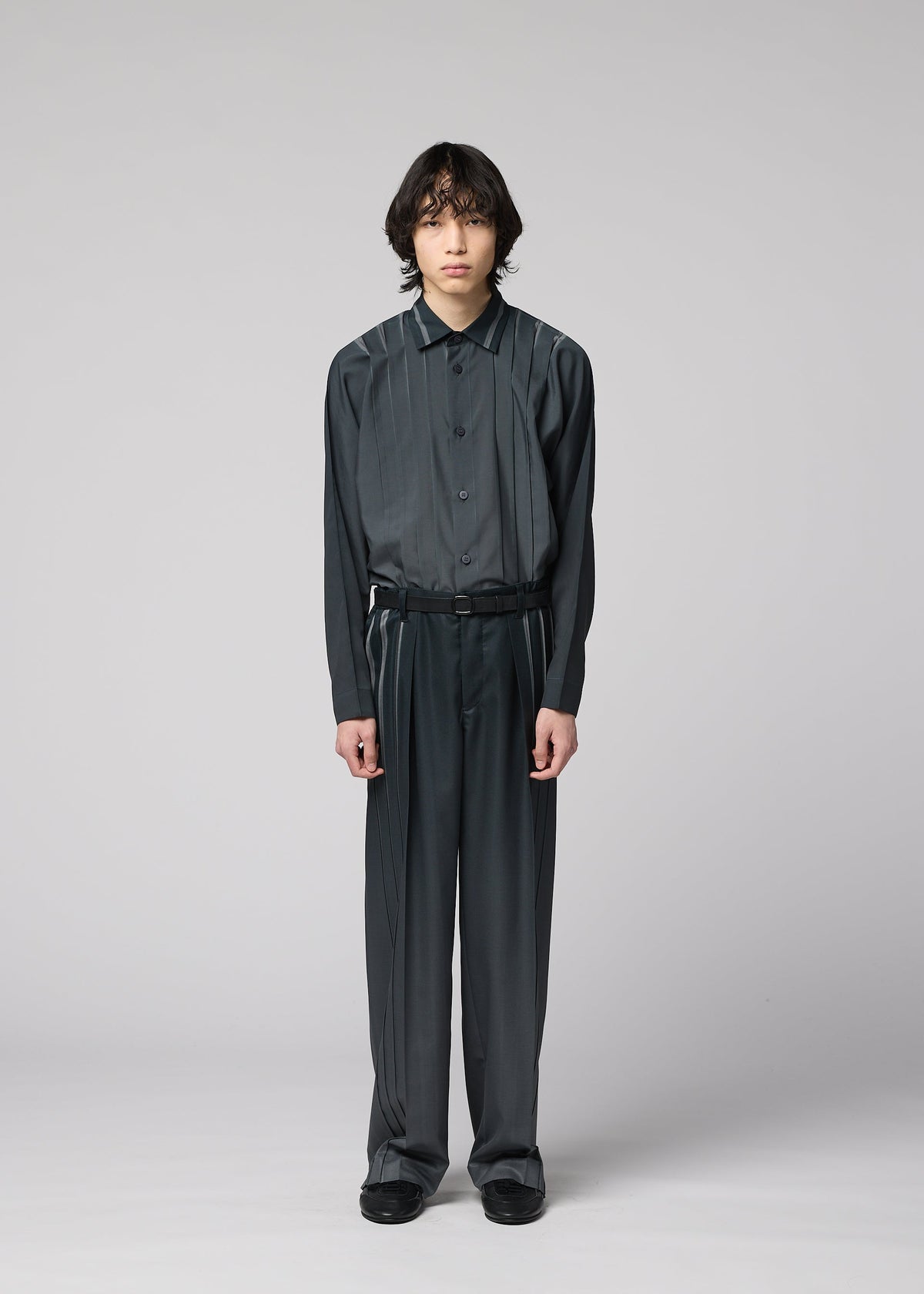 A person with medium-length dark hair stands against a plain gray background, wearing the DAWN RAY dark, loose-fitting button-up shirt and matching pleated pants from IM MEN, which feature a subtle gradation pattern, paired with black shoes. view 4 of 7